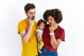 Interracial young family of black mother and hispanic father with daughter pointing to the eye watching you gesture, suspicious expression