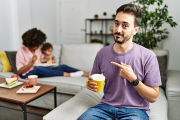 Hispanic father of interracial family drinking a cup coffee pointing with hand finger to the side showing advertisement, serious and calm face