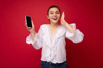 Attractive young happy brunet woman wearing white blouse standing isolated over red background showing mobile phone with empty screen for cutout looking at camera and announcing
