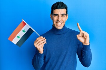Handsome hispanic man holding syria flag smiling with an idea or question pointing finger with happy face, number one