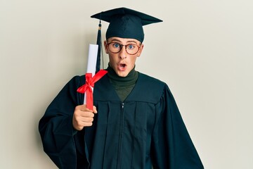 Young caucasian man wearing graduation cap and ceremony robe holding diploma scared and amazed with...
