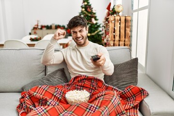 Young hispanic man eating popcorn sitting on sofa by christmas tree annoyed and frustrated shouting with anger, yelling crazy with anger and hand raised