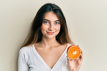 Young caucasian woman holding fresh slice of orange looking positive and happy standing and smiling...