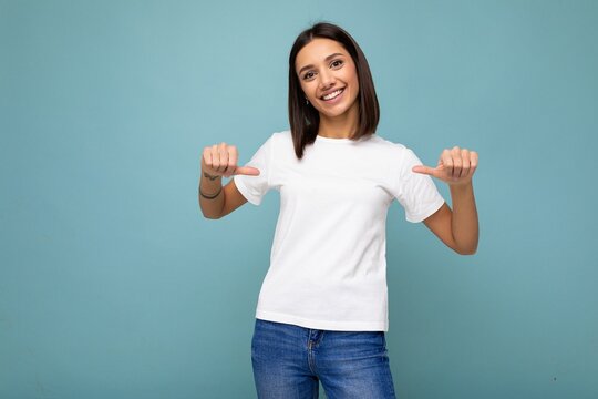 Portrait of positive happy smiling young pretty cute brunette woman with sincere emotions wearing casual white t-shirt for mockup isolated over blue background with copy space and pointing at empty