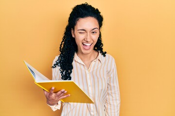 Young hispanic woman with curly hair holding book winking looking at the camera with sexy...