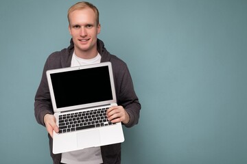 Close-up portrait of handsome blonde man holding computer laptop looking at camera isolated over blue background