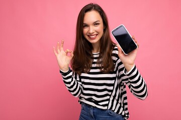 beautiful happy young woman wearing striped sweater isolated over background with copy space showing ok gesture looking at camera showing mobile phone screen display