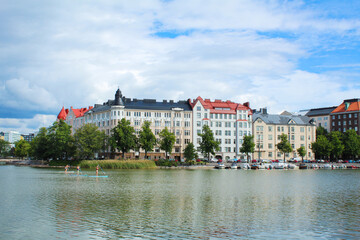 Fototapeta premium Beautiful building on river bank and green trees under blue sky with clouds, people sailing on paddleboards 