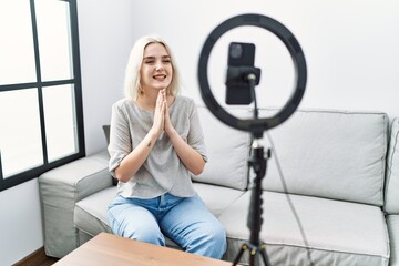 Fototapeta na wymiar Young caucasian woman recording vlog tutorial with smartphone at home praying with hands together asking for forgiveness smiling confident.