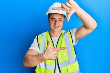 Handsome young man wearing safety helmet and reflective jacket smiling making frame with hands and fingers with happy face. creativity and photography concept.