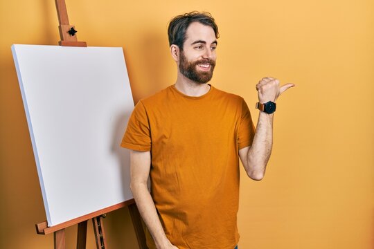 Caucasian man with beard standing by painter easel stand pointing thumb up to the side smiling happy with open mouth