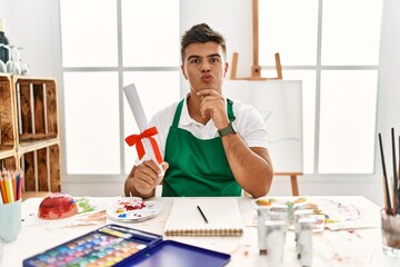 Young hispanic man at art studio holding degree looking at the camera blowing a kiss on air being lovely and sexy. love expression.