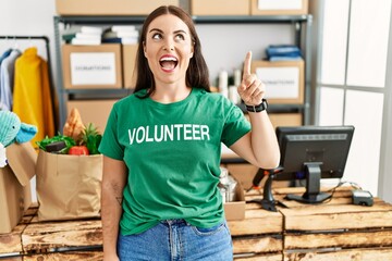 Young brunette woman wearing volunteer t shirt at donations stand pointing finger up with...