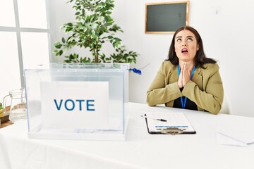 Young brunette woman sitting at election table with voting ballot begging and praying with hands...
