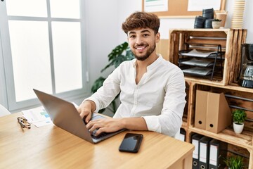 Young arab man working using computer laptop at the office looking positive and happy standing and smiling with a confident smile showing teeth