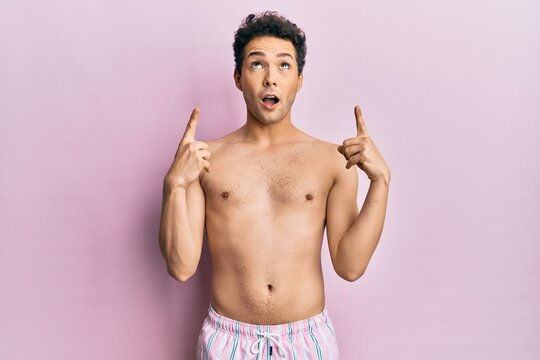 Young handsome man wearing swimwear shirtless amazed and surprised looking up and pointing with fingers and raised arms.
