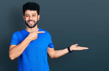 Young arab man with beard wearing casual blue t shirt amazed and smiling to the camera while presenting with hand and pointing with finger.