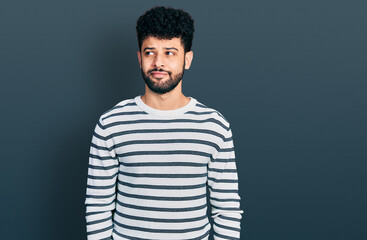 Young arab man with beard wearing casual striped sweater smiling looking to the side and staring away thinking.