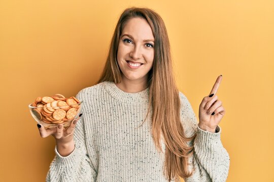 Young blonde woman holding salty biscuits bowl smiling happy pointing with hand and finger to the side