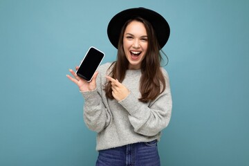 Closeup of winking charming young happy woman wearing black hat and grey sweater holding phone...