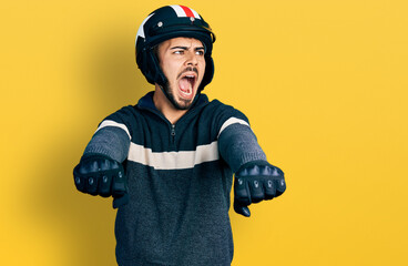 Young hispanic man with beard wearing motorcycle helmet doing driving gesture angry and mad...