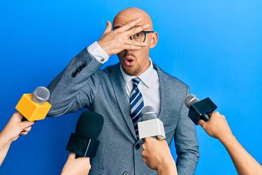 Bald man with beard being interviewed by reporters holding microphones peeking in shock covering face and eyes with hand, looking through fingers afraid