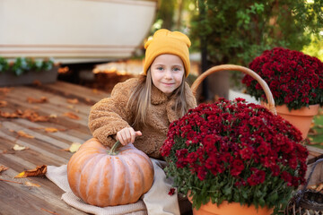 Happy little girl sitting on porch of house with chrysanthemums potted and pumpkins. home fall...