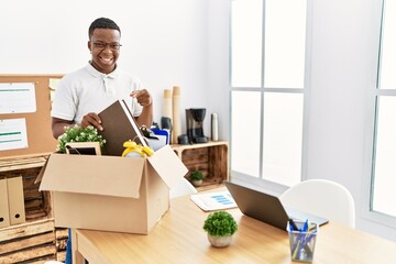 Obraz na płótnie Canvas Young african man putting office objects into cardboard box smiling happy pointing with hand and finger