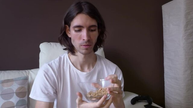 Young man eating peanuts out of a glass bowl while watching comedy series