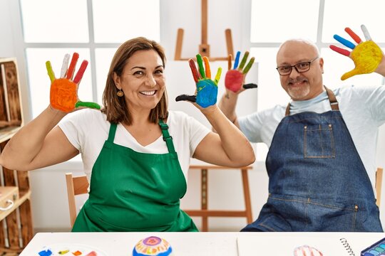 Middle age hispanic painter couple smiling happy showing painted palm hands at art studio.
