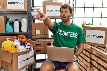 Young handsome man wearing volunteer t shirt using laptop pointing with finger surprised ahead,...