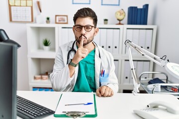 Young man with beard wearing doctor uniform and stethoscope at the clinic asking to be quiet with finger on lips. silence and secret concept.