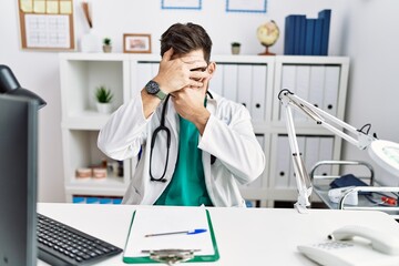 Young man with beard wearing doctor uniform and stethoscope at the clinic covering eyes and mouth with hands, surprised and shocked. hiding emotion