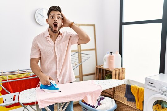 Young man with beard ironing clothes at home crazy and scared with hands on head, afraid and surprised of shock with open mouth