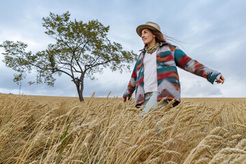 Happy woman in a hat and a bright shirt on a wheat field on an autumn day. In the background, a lonely tree against the sky with clouds.