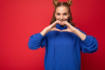 Young happy positive attractive blonde woman with two horns with sincere emotions wearing stylish bright blue hoodie isolated on red background with copy space and showing heart gesture