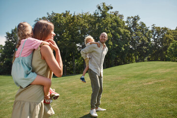 Little kids enjoying piggyback ride on the back of their mom and dad. Family walking in green...
