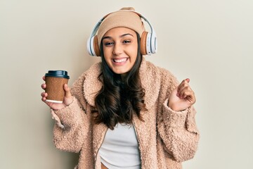 Young hispanic woman using headphones drinking coffee screaming proud, celebrating victory and success very excited with raised arm
