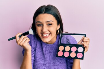 Young hispanic woman holding makeup brush and blush smiling and laughing hard out loud because...