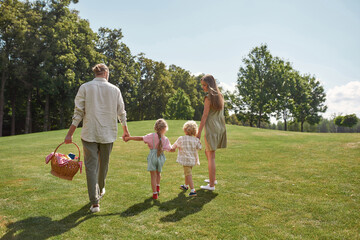 Obraz premium Back view of young family of four holding hands together while walking in green park. Family having picnic in nature on a summer day