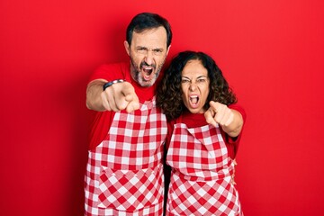 Middle age couple of hispanic woman and man wearing cook apron pointing displeased and frustrated to the camera, angry and furious with you