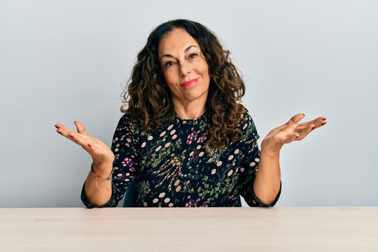 Beautiful middle age woman wearing casual clothes sitting on the table clueless and confused expression with arms and hands raised. doubt concept.