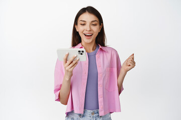 Young modern woman watching on mobile phone, looking at smartphone with cheerful smiling face expression, white background