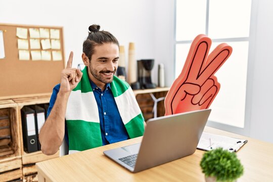 Young hispanic man watching football on laptop cheering game at the office surprised with an idea or question pointing finger with happy face, number one
