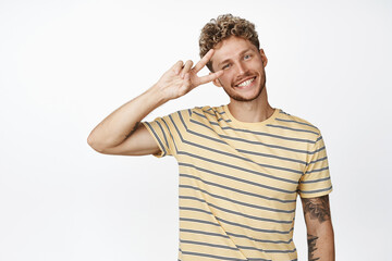 Fototapeta premium Happy smiling blond man shows peace sign salute and looking at camera positive, standing over white background