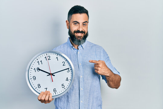 Young hispanic man holding big clock smiling happy pointing with hand and finger