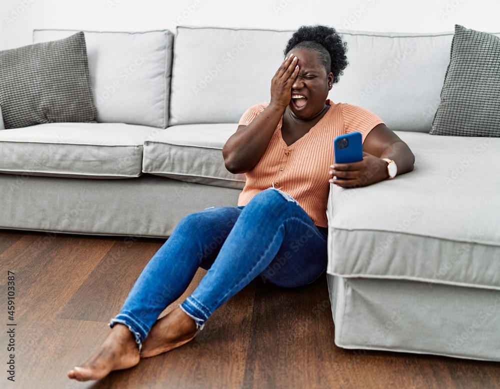 Wall mural Young african woman using smartphone sitting on the floor at home yawning tired covering half face, eye and mouth with hand. face hurts in pain.