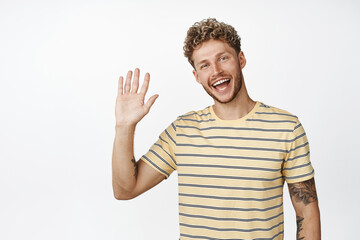 Friendly blond guy saying hello, waving hand and smiling at camera, standing over white background