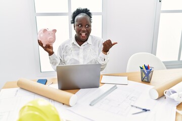 Young african woman working at architecture studio holding piggy bank pointing thumb up to the side smiling happy with open mouth