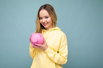 Portrait photo of happy positive smiling young beautiful attractive blonde woman with sincere emotions wearing trendy yellow hoodie isolated over blue background with free space and holding pink pig